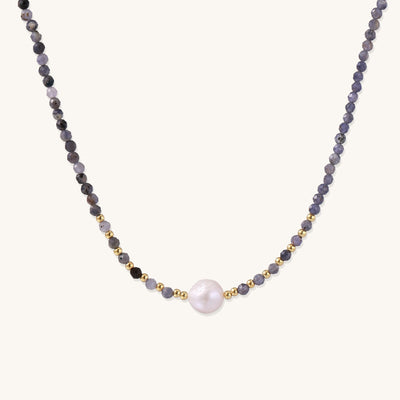 Delicate beaded necklace with violet iolite stones, gold spacers, and central white freshwater pearl on neutral background.
