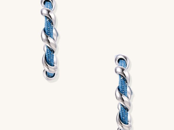 Twisted hoop earrings featuring blue denim fabric braided with gold-plated metal in a spiral design on white background.