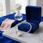 A silver cable bracelet with a heart charm engraved with a heart, displayed in front of an open dark blue velvet jewelry box on a marble slab. A folded blue velvet pouch and a white flower in a glass vase are visible in the background.