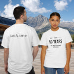 Two models wearing white graphic t-shirts in front of a mountain backdrop. The model on the left shows the back of the shirt with 'LastName' printed in black, and the model on the right shows the front of the shirt with 'AUTHENTIC 50 YEARS OF NOT GIVING A DAMN' printed in black.