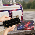 A black zip-around wallet with gold-tone hardware is shown open in hands, with a matching long wallet featuring a red, blue, and purple galaxy print placed beside it. A matching galaxy-print bag is also visible in the background on a wooden table outdoors.