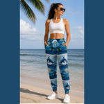 A woman stands on a beach wearing blue patterned jogger pants with a dolphin print, a white crop top, and white sneakers. The background shows the ocean, sand, and a palm tree.