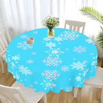 Round table with a blue tablecloth featuring white snowflakes, surrounded by white chairs in a room with light-colored curtains.