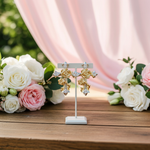 Gold and silver floral earrings on a stand with flowers in the background
