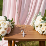 Silver cross necklace on a wooden table with floral arrangements against a pink curtain.