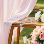 Silver cross necklace on a wooden table with pink and white flowers and pink fabric in the background.
