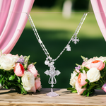 Silver cross necklace over a stand with pink and white flowers in the background